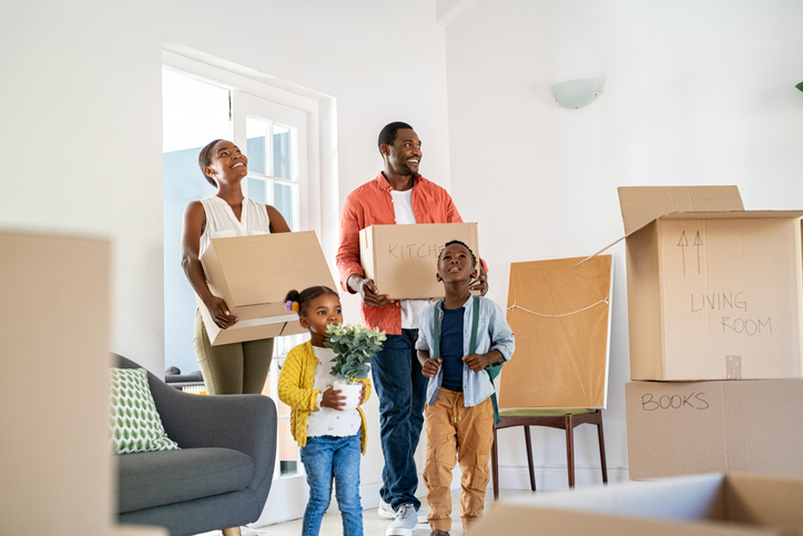 A family with two children carrying boxes in a new home. Cheerful mother and father holding boxes while entering new home with son and daughter. Happy son and daughter helping parents relocating in new house.